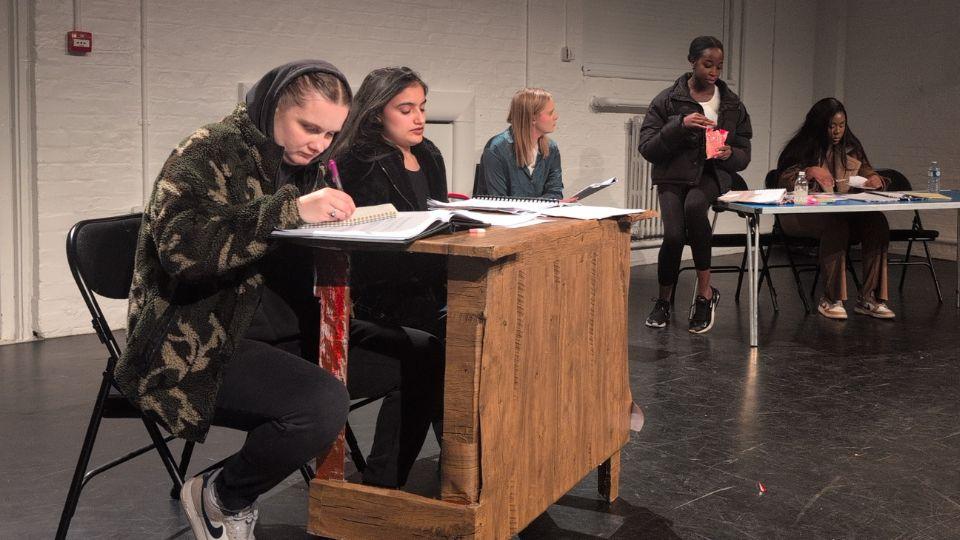 Five young females sit on and around desks, looking at paperwork, learning materials