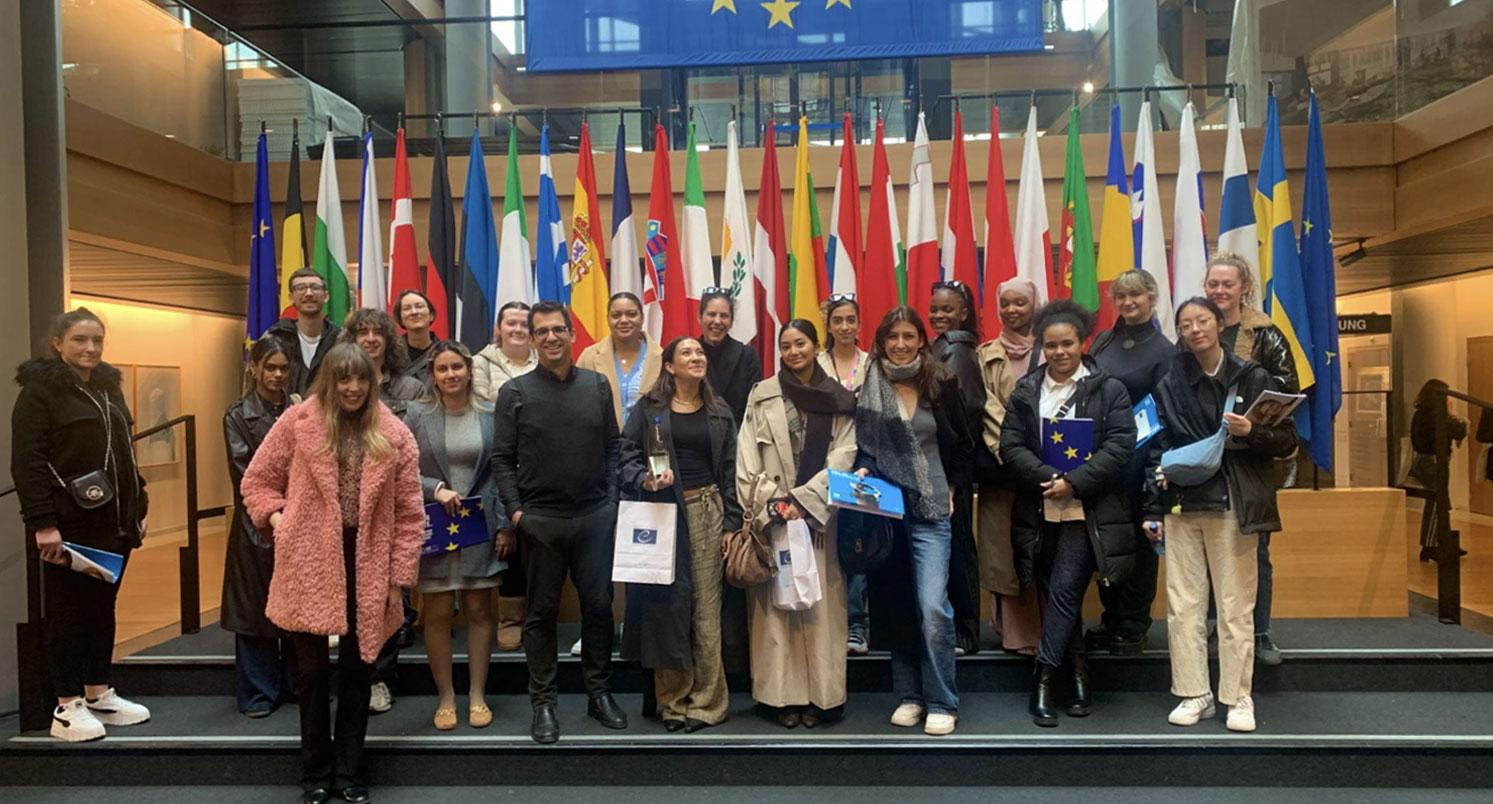 Twenty law students standing on a step in front of a collection of European flags at the European Parliament