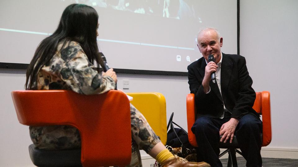 Leah Borromeo sits opposite Adam Curtis, who is wearing a black jacket and speaking into a microphone. 