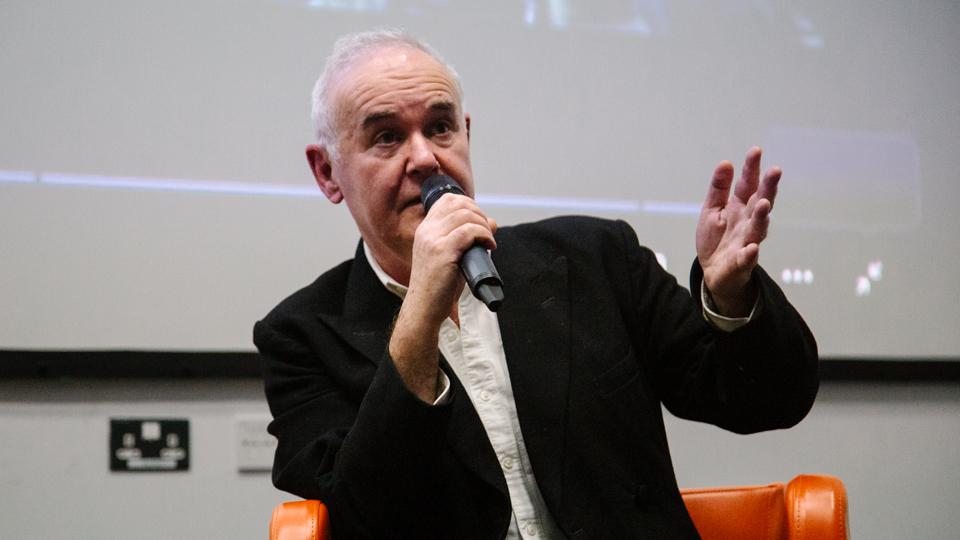Adam Curtis wearing a black jacket sits in a chair and speaks into a microphone. 