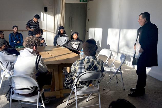 Students sit in a brightly lit classroom, listening to a man speak
