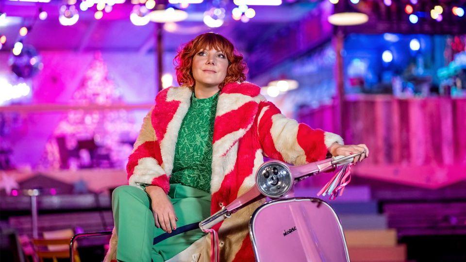 Sophie Willan as Alma wearing green and a pink and white coat, sitting on a pink scooter