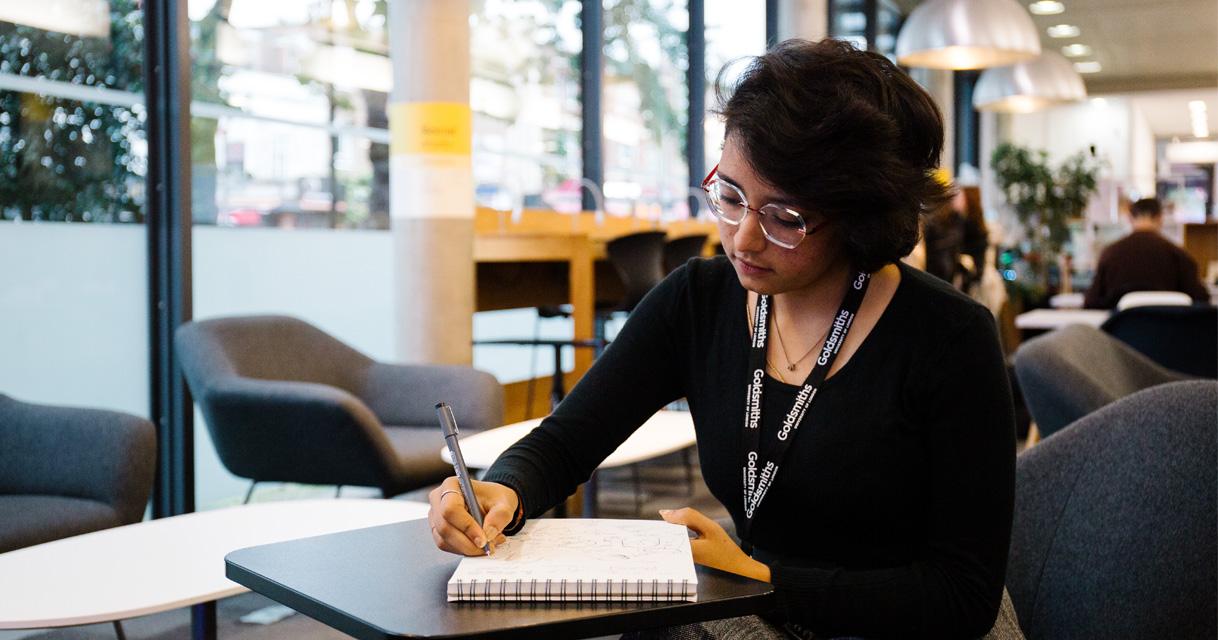A student writes in a notebook in a modern university library.