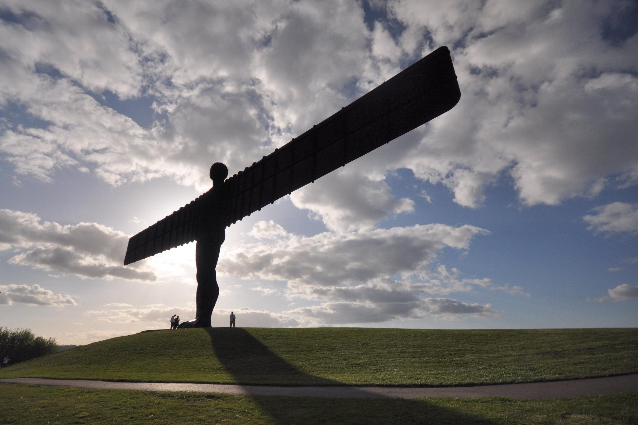 A photo showing the silhouette of the Angel of the North, against a cloudy sky with people at the foot of the sculpture. 