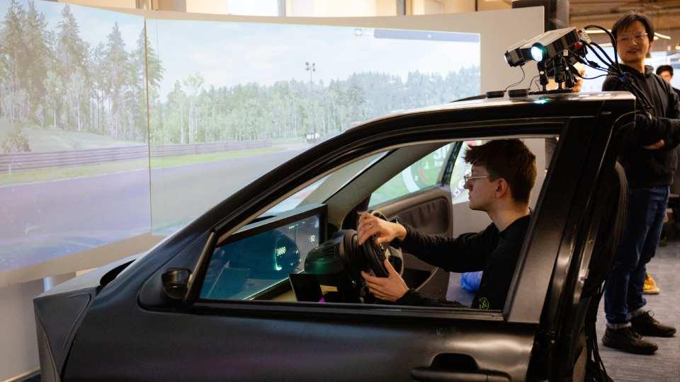 Male sites in front seat of car, hands on steering wheel, looking through windscreen to digitised landscape
