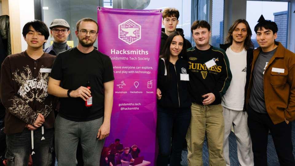 Group of young people pose for picture, huddling around a pink sign that reads Hacksmiths Society./ 
