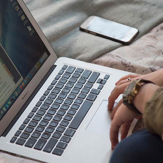 Close up of a laptop on a bed with a phone next to it.