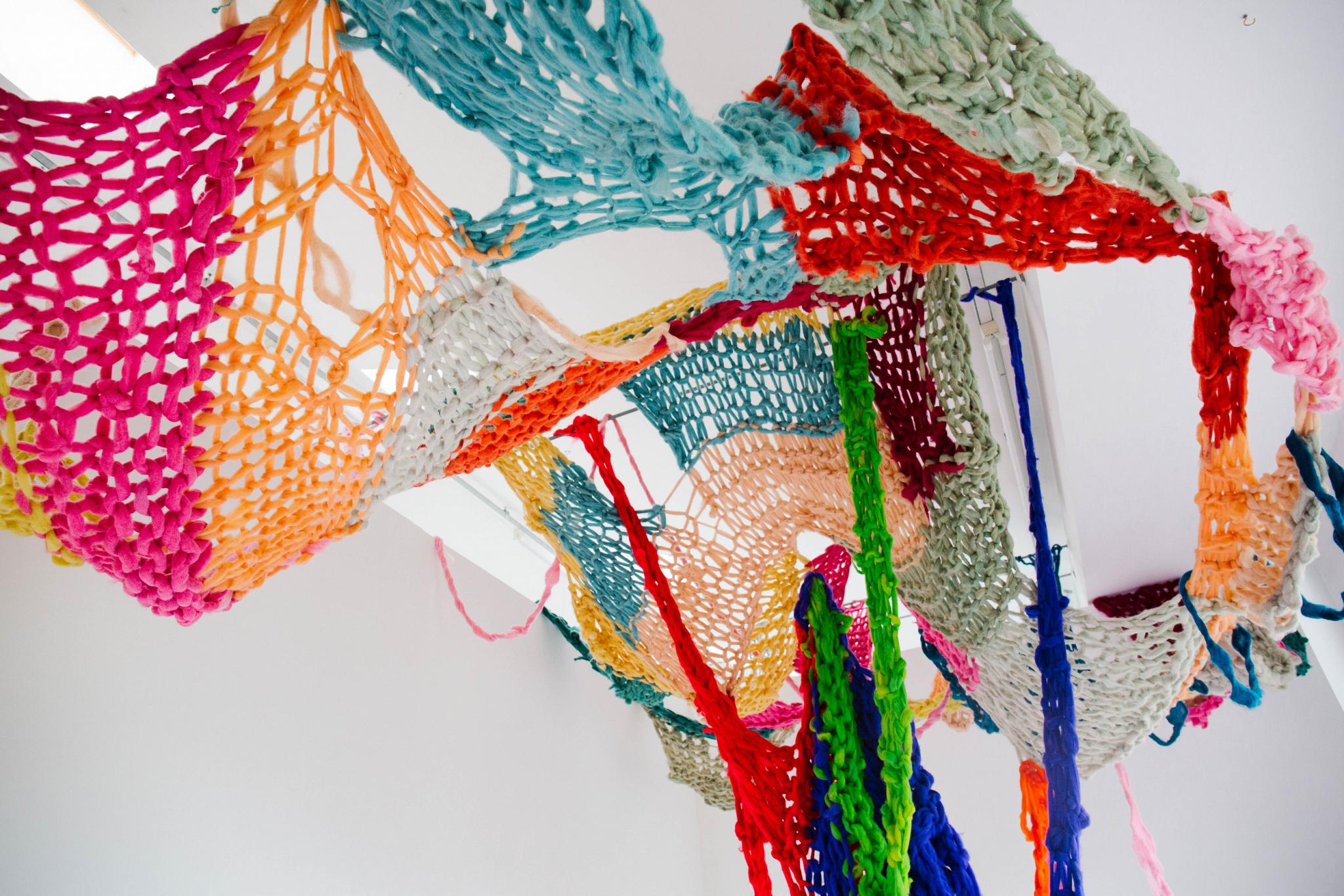 Colourful, hand-knitted net-like textile installation suspended from the ceiling in a gallery space