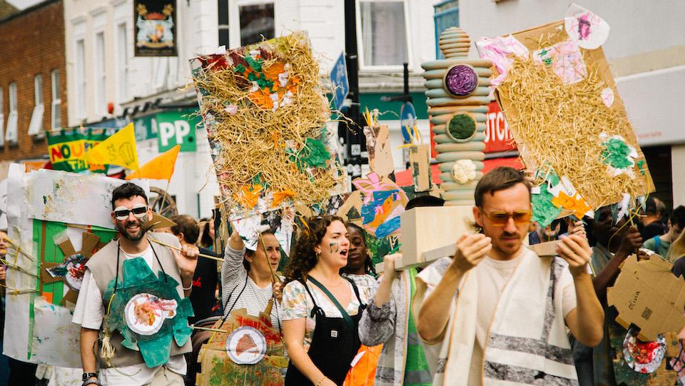 A group of people walking as part of a parade during Deptford X, wearing handmade costumes and holding handmade banners.