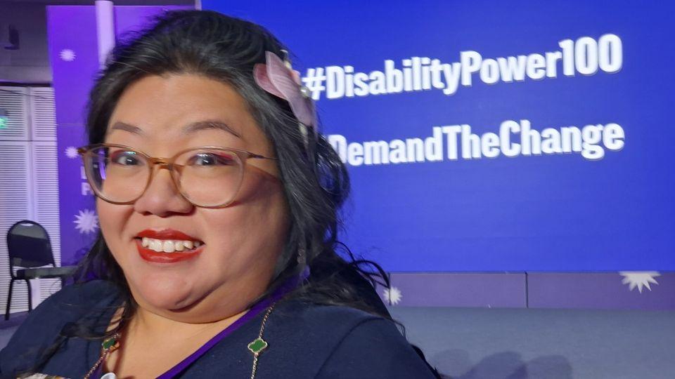 Dr Carmen Yau is smiling in front of a blue screen which says #DisabilityPower100 #DemandTheChange 