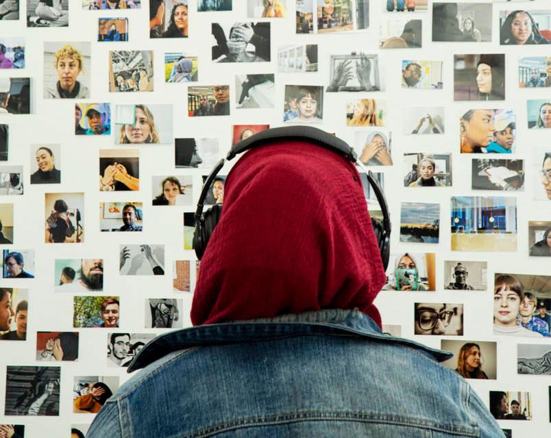 A woman wearing a headscarf and headphones looks at a wall of pictures.