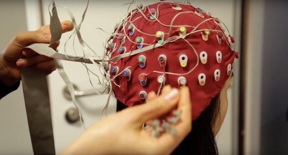 Hands are shown fittings probes to someones head as part of a psychology experiment.  