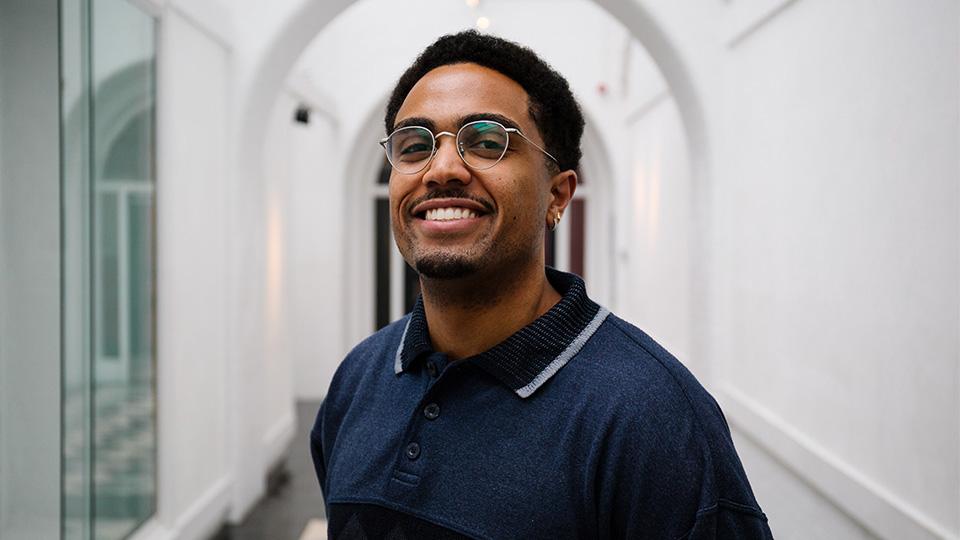A portrait of Alex, a Black man, smiling in a white corridor