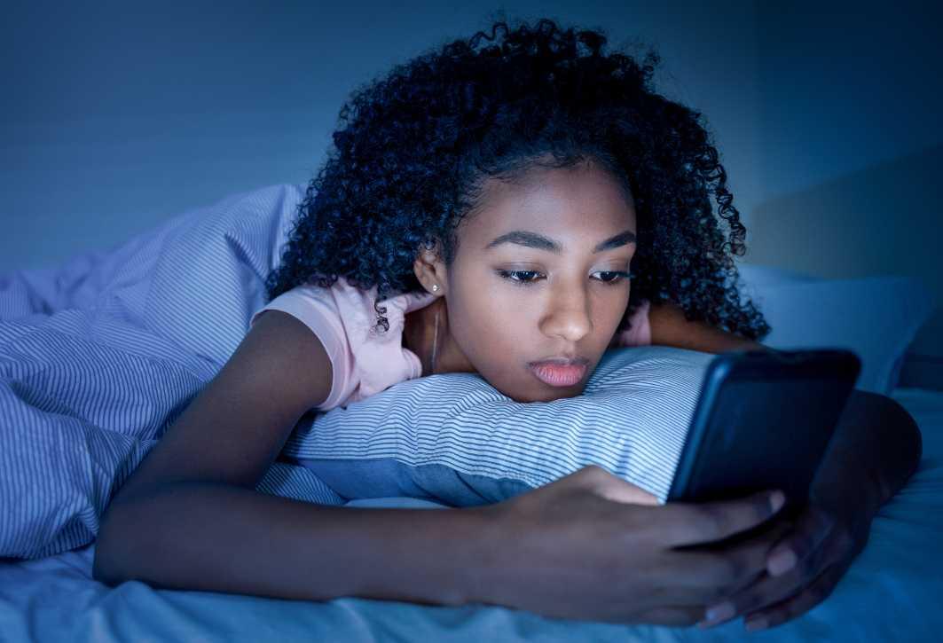 A sad-looking girl looks at her smartphone while lying in bed