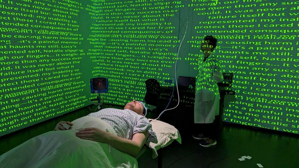 A person lying on a hospital bed with another person in a lab coat standing nearby and green text projected onto the walls