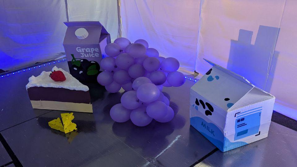 Oversized props for 'The Golden Cheese' including giant cartons, giant grapes made from purple balloons and a giant piece of cake