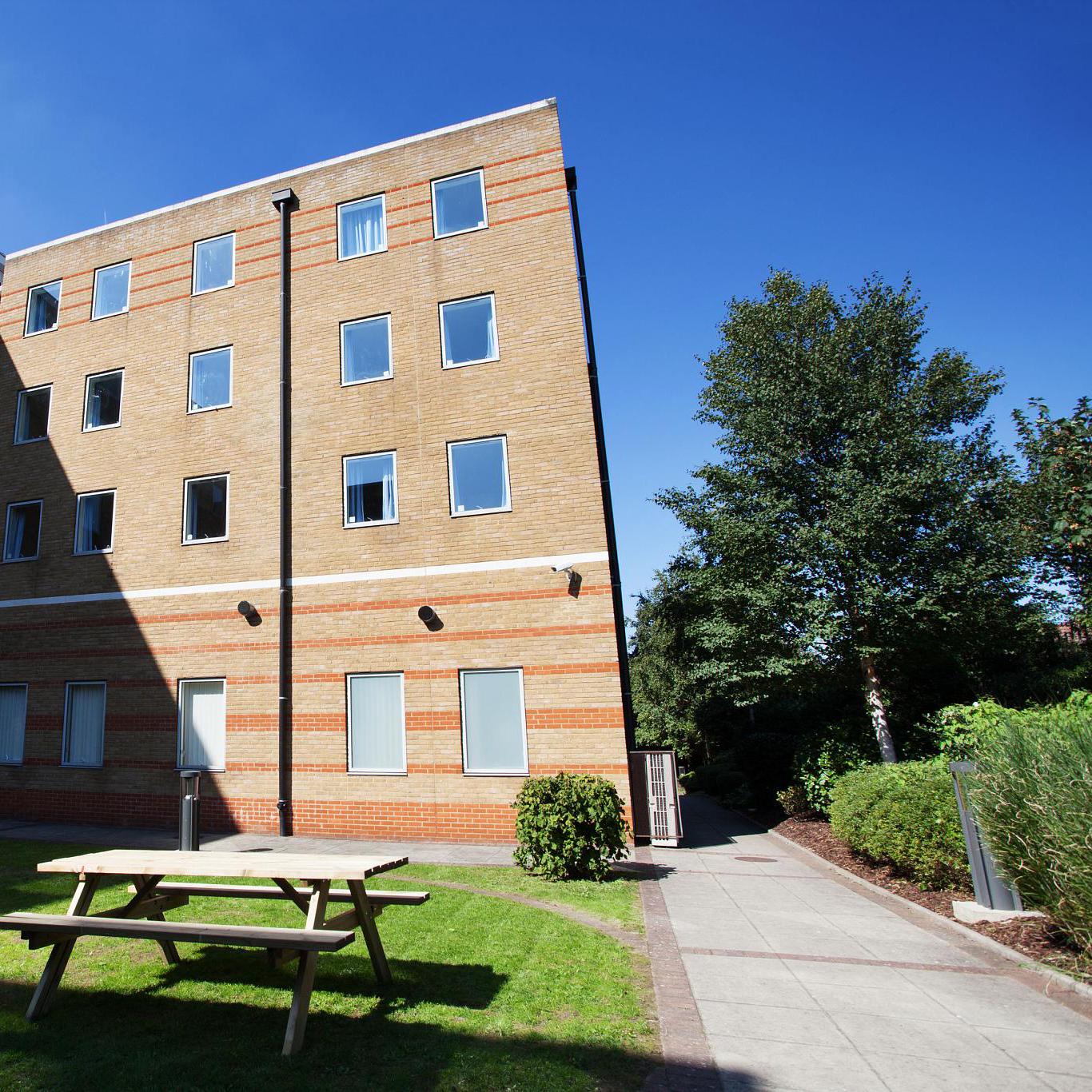 Outside of a brick small tower block with a bench outside on a sunny day.