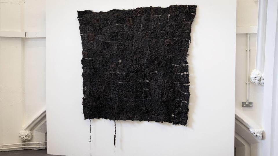 A black woven artwork hanging in a white exhibition space