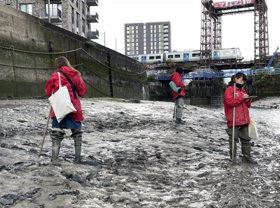 Three students stand making observational notes at Deptford Creek.