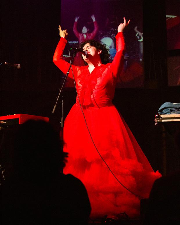 A woman in a red dress sings on a stage with her arms up