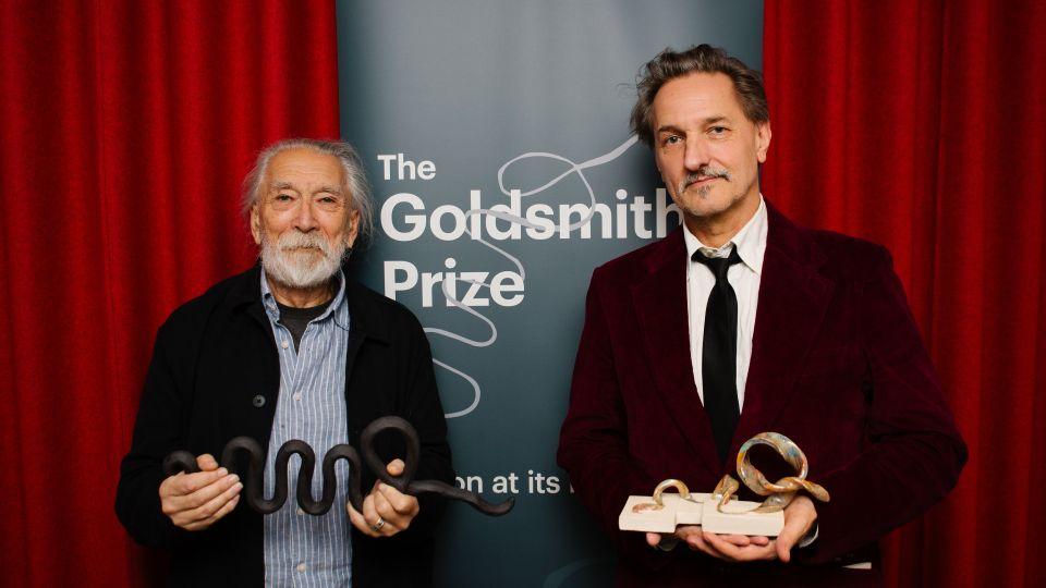 M John Harrison holding his trophy and C.D. Rose holding his ceramic trophy in front of a banner that says The Goldsmiths Prize and a background of red curtains 