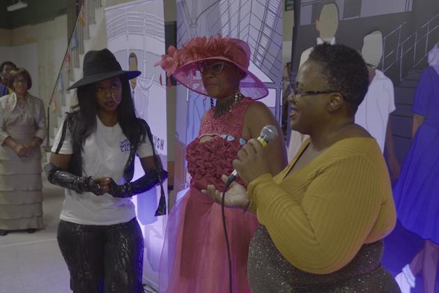Three women speaking to an audience at an event. The woman to the right holds the microphone, whilst the woman at the centre of the image is dressed in a vibrant pink dress