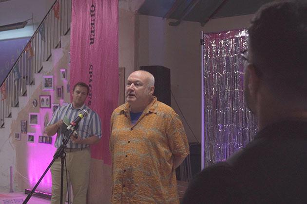 A man stands in front of a purple and pink tinsel background, speaking into a microphone whilst a crowd watches.