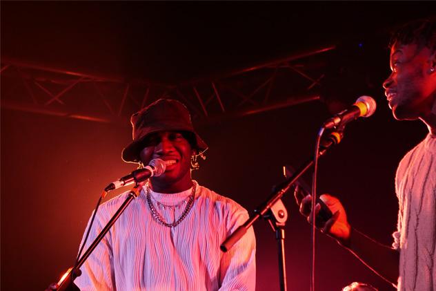 Two men perform on stage, they are lit in red and are behind standing microphones