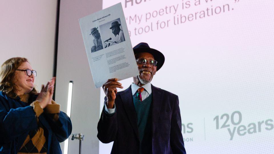 Female (left) smiles as she hand plaque to male in hat (also smiling, right)