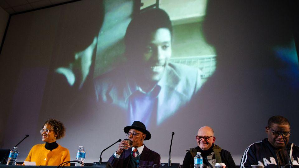 one female and three males sit behind table with a backdrop of young black man (LKJ) behind them. 
