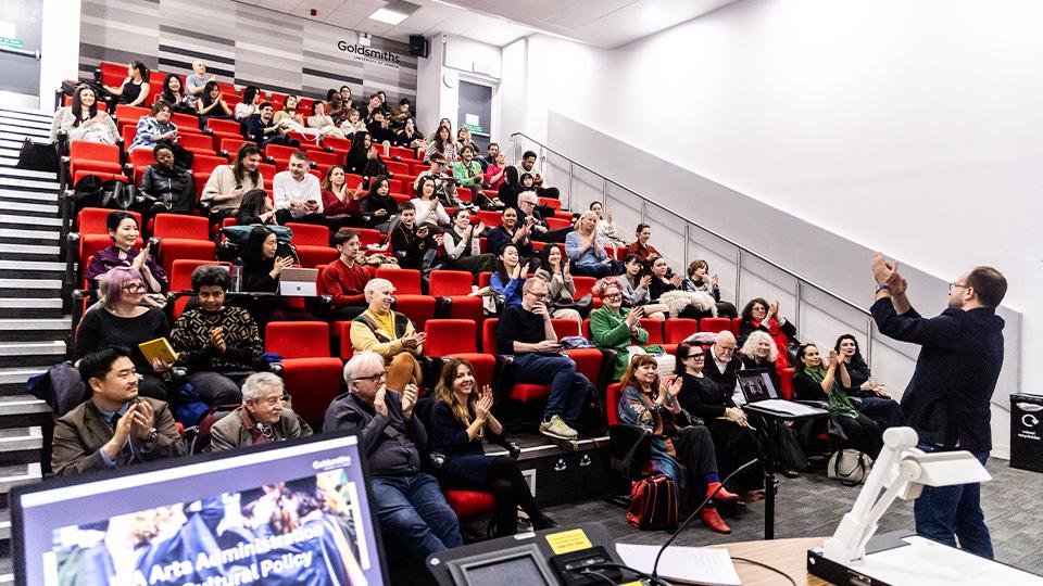 Academic Dr Aleksander Brkić stands at the front of a lecture theatre, applauding attendees sitting on raked seats. 