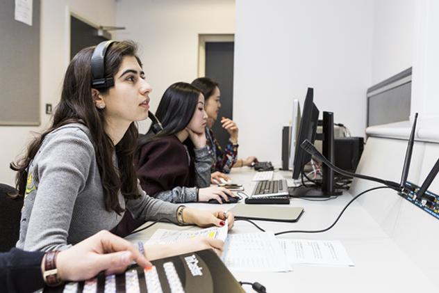 Three students sit at the gallery that forms part of the TV studio &ndash; the student in the foreground wears a headphone and mic and holds a script in their hand