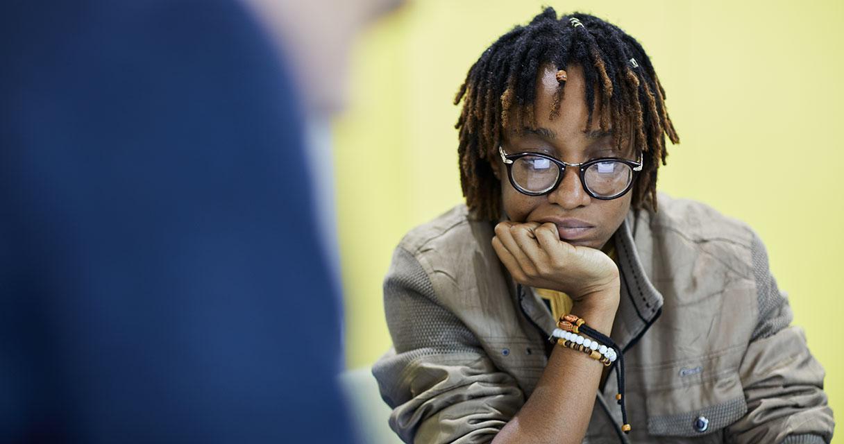 A photograph of a woman sitting across from a man with a yellow wall behind her. The man is in the foreground of the left-hand side of the image.