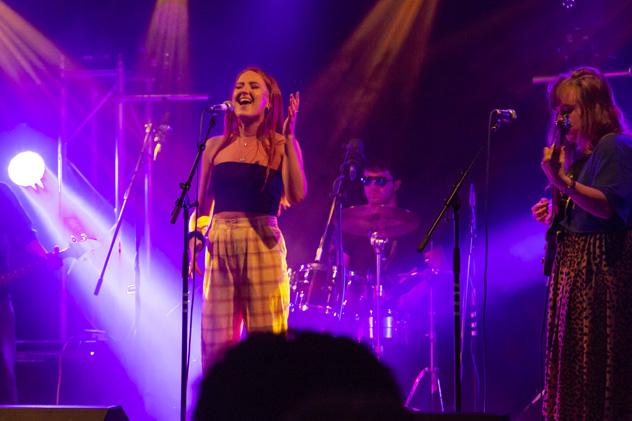 A music student stands in front of a microphone, singing. There are two bandmates in the background &ndash; one on drums, and one playing a guitar.