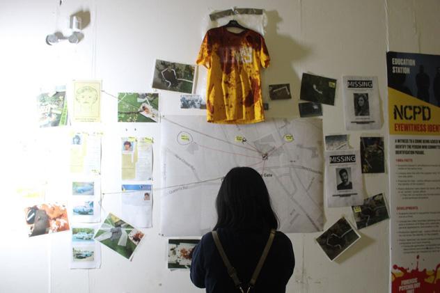 A person looking at a wall containing evidence from a crime scene, including a bloodied t-shirt and a map