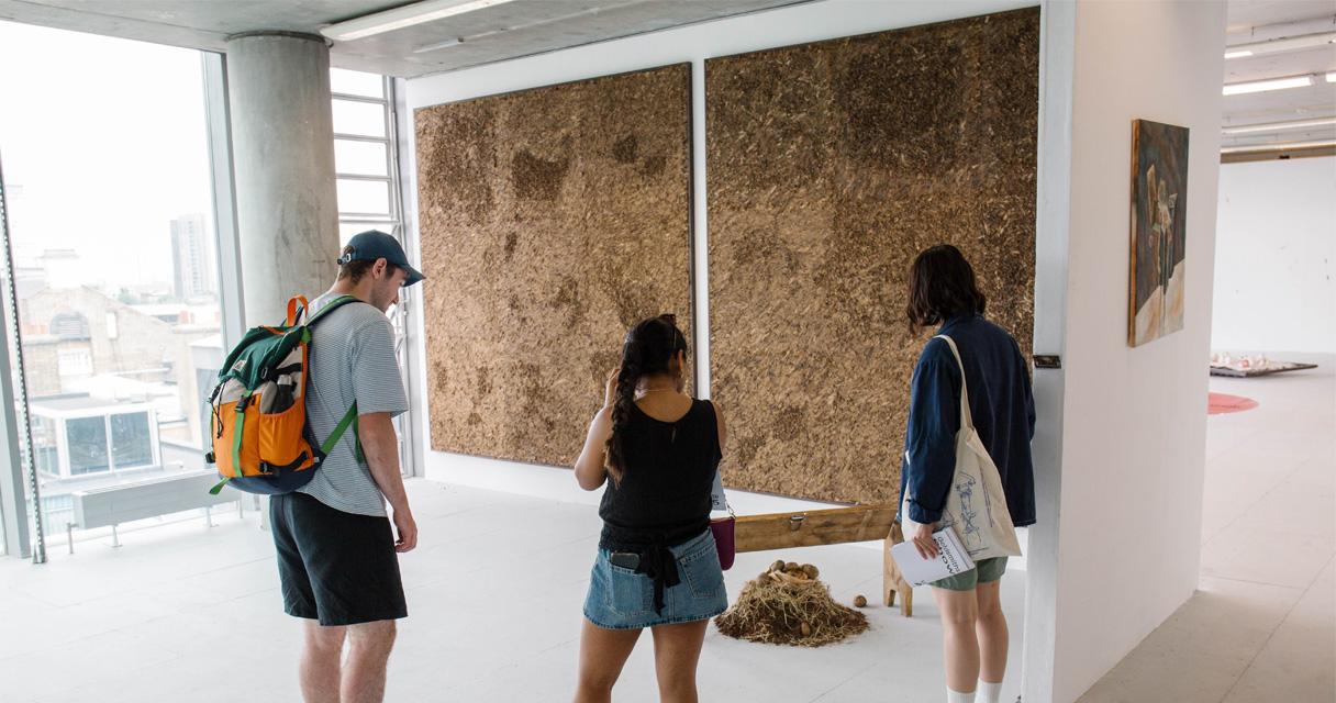 People look at an installation of small organic mounds in a large exhibition space which looks out over the London skyline