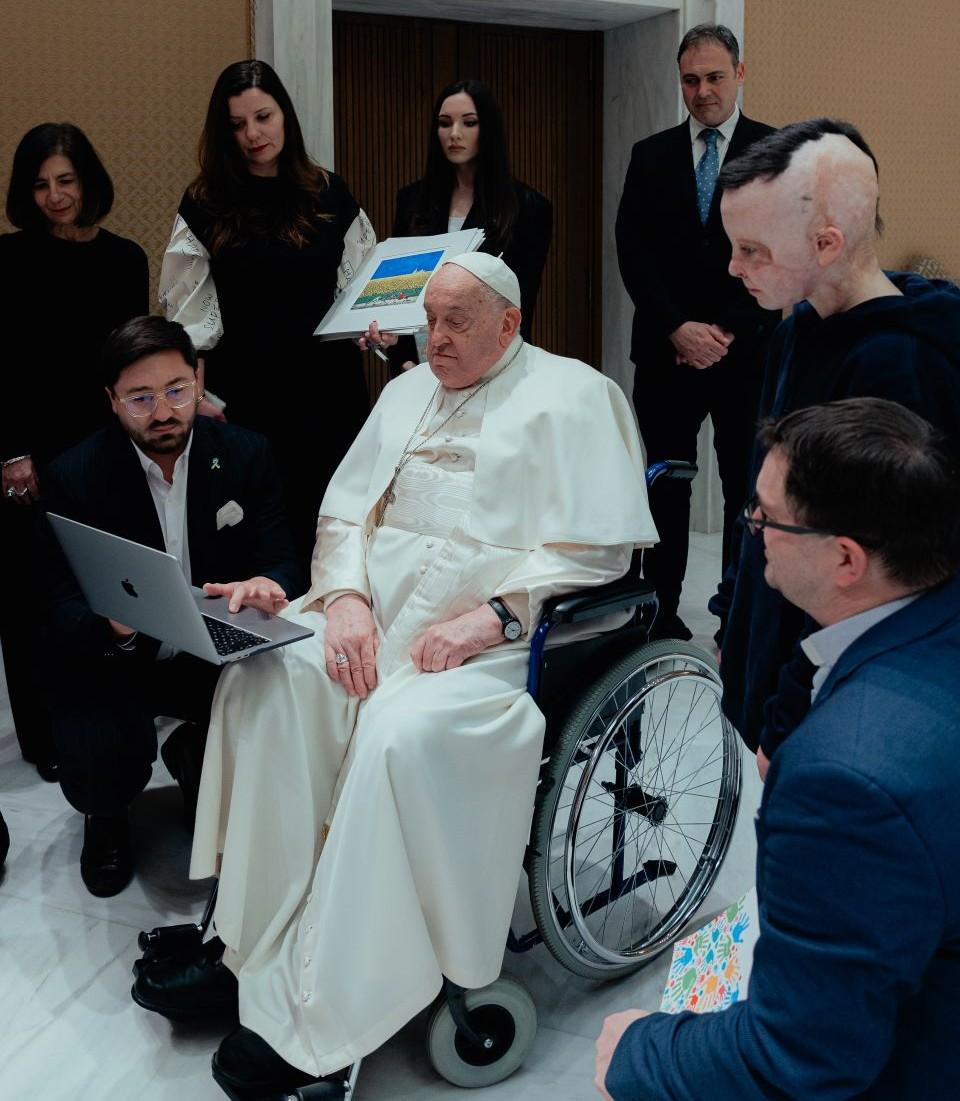 Pope Francis watching the film on a laptop, surrounded by people. 