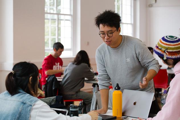 Several postgraduate students sit and stand around a desk in a seminar, chatting to each other