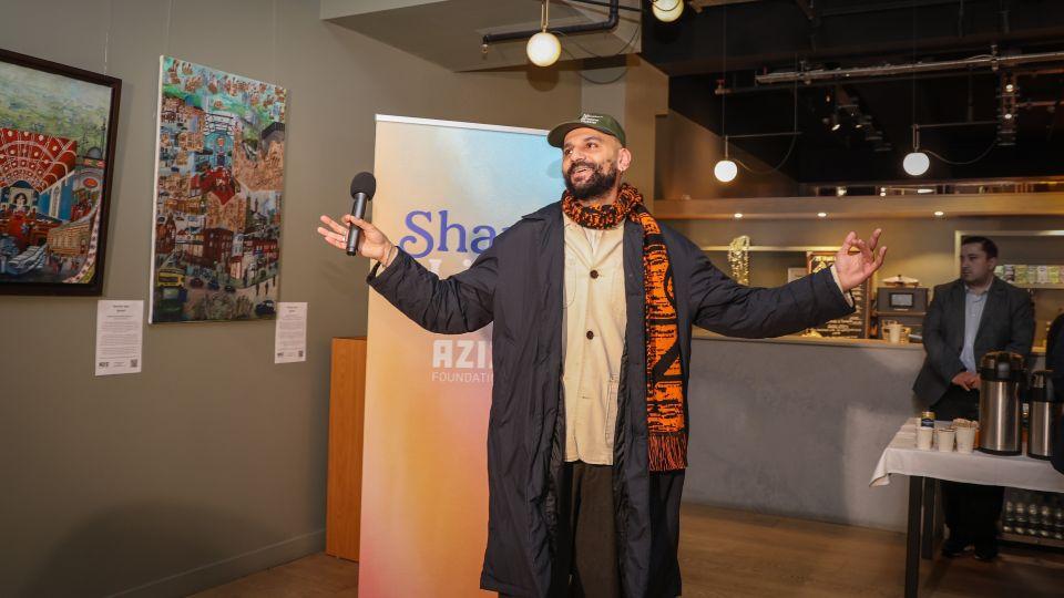 Raheel Khan stands in front of some of the artwork, holding a microphone and giving a speech at the opening of the exhibition