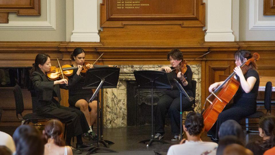 A string quartet performing in front of a wood-panelled wall while an audience looks on. 