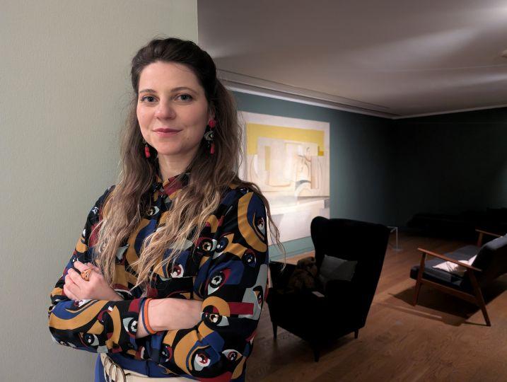 Sasha, wearing a colourful printed shirt, stands in a room with dark green walls, with a sofa and an arm chair. 
