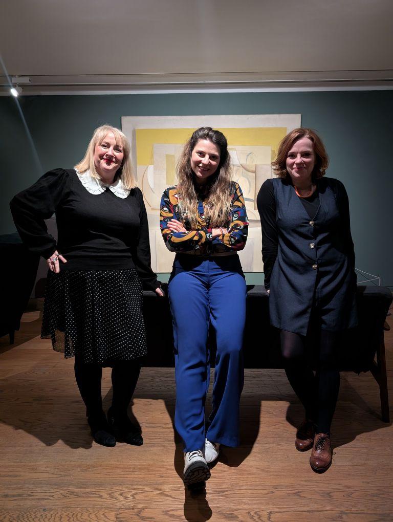 Three people stand in front of a sofa in a room with dark green walls and a large painting is hanging behind them. 