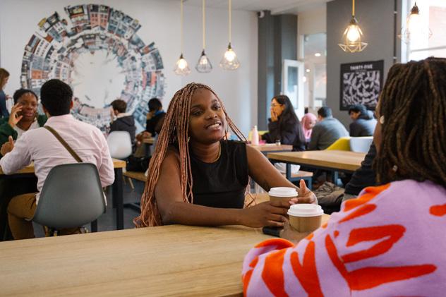 Two students on a long table chat together over hot drinks, with other people in the cafe in the background