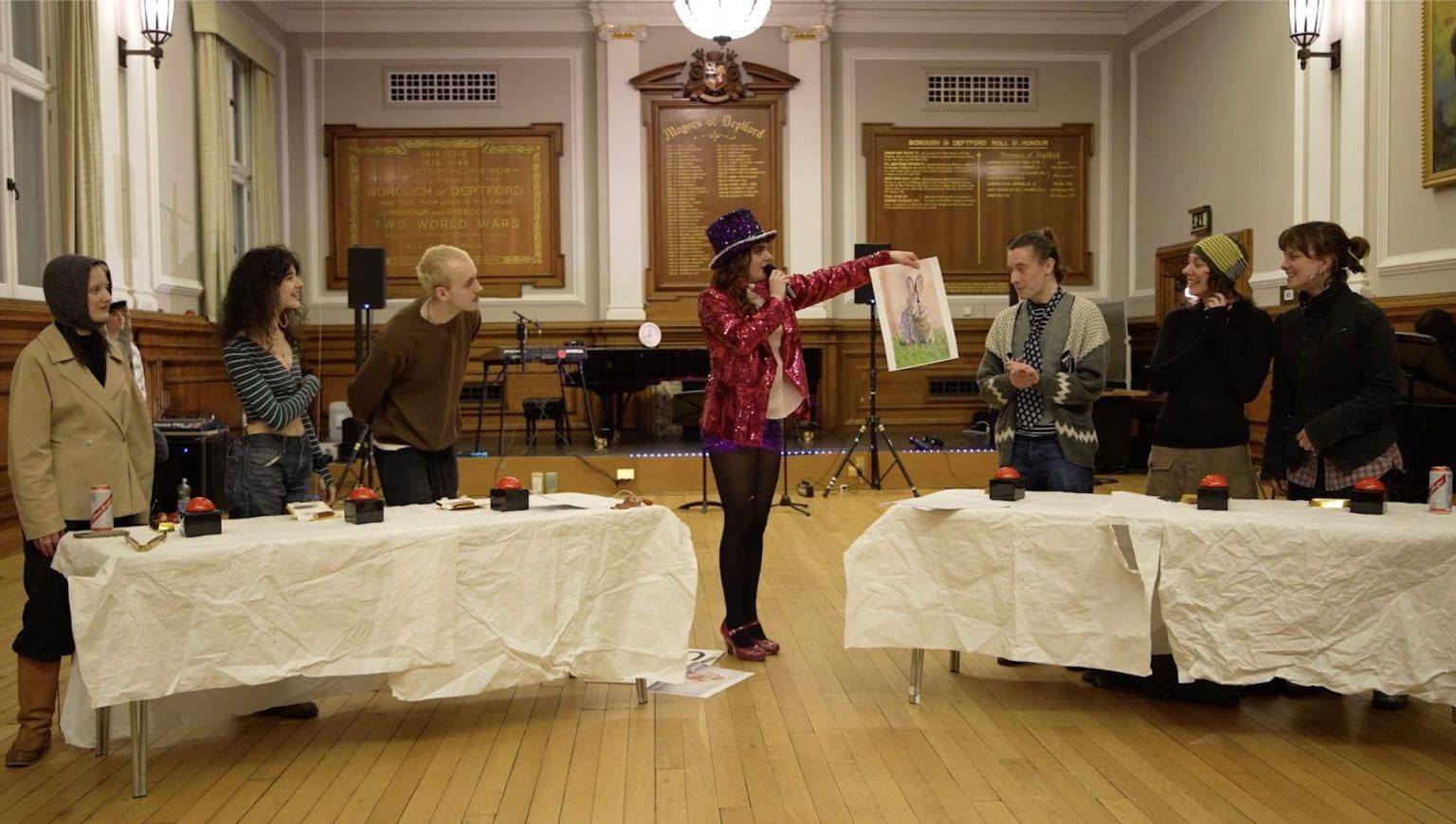 Female Performer in top hat, sequinned jacket and high heels MCs panel of guests