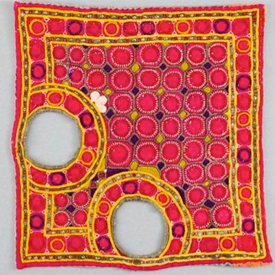 andari - A square of fairly course red fabric with two large holes half way along two of the edges at right angles. The textile is layered and embroid