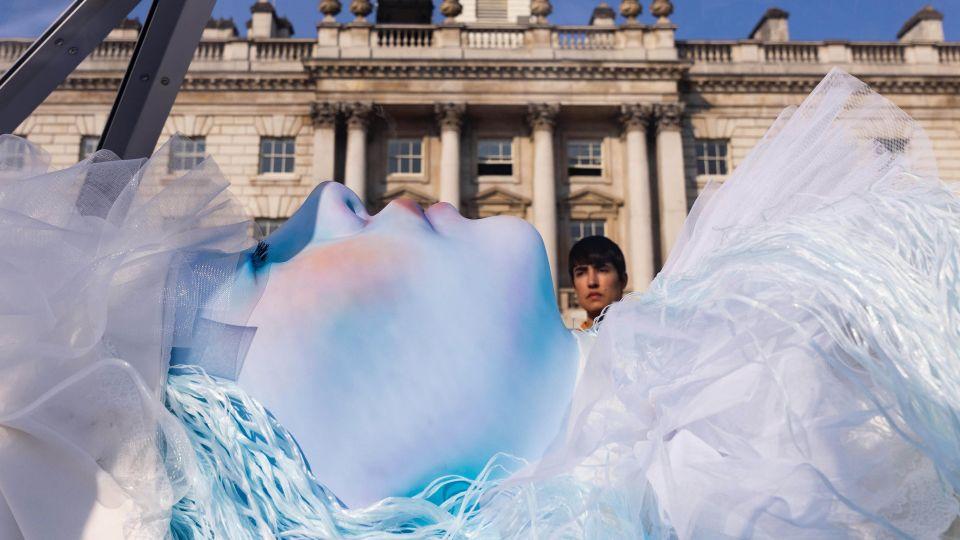 Blue statue-like female in repose against backdrop of Somerset House. 