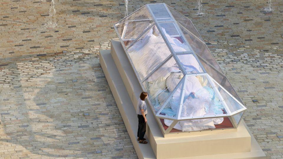 A giant blue figure in a glass casket, with a visitor looking in