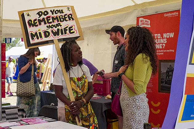 Two women in conversation. The woman on the left holds a placard that reads 'Do you remember your midwife?'
