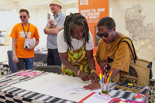 Two women in conversation whilst leaning over a white piece of paper. On the paper the woman on the left draws around the hand of the woman on the right.