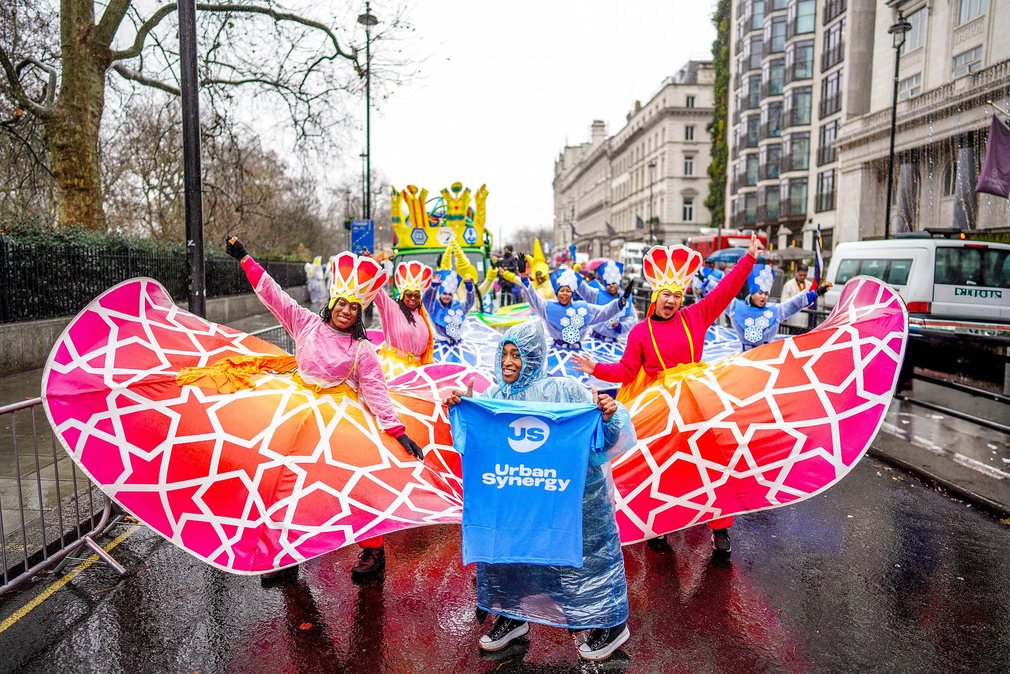 Performers in the parade in colourful costumes, with a person stood in front holding an Urban Synergy T-shirt